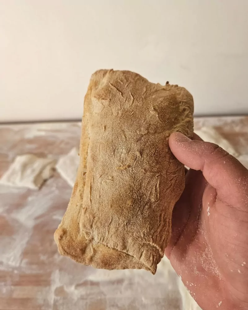 Ciabatta artesanal enharinada sostenida en la mano sobre mesa de trabajo, Bona Biga Pan Mendoza